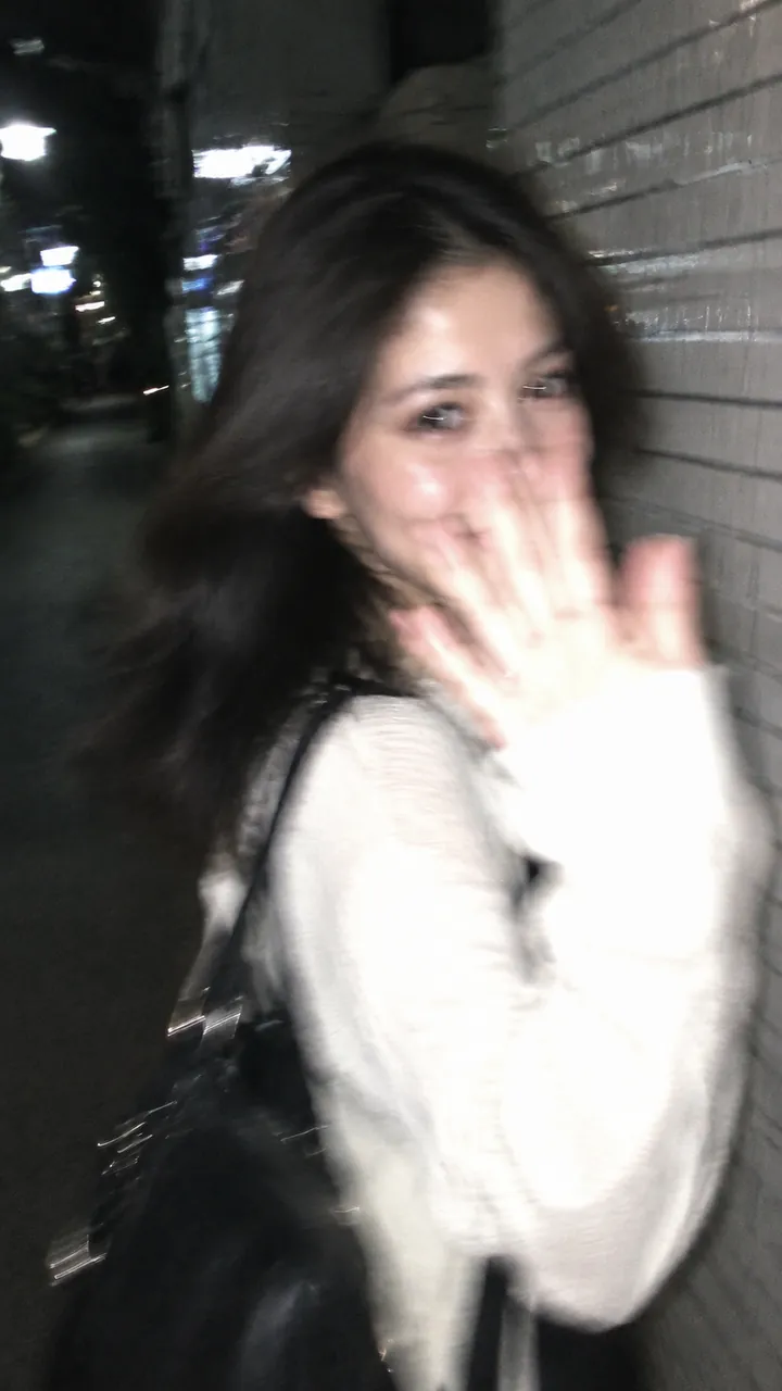 Blurry night smartphone snapshot of a dark-haired woman in a white jacket raising her hand toward the lens beside a white-tiled wall