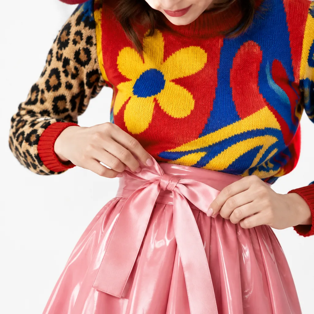 Close-up of hands tying a pink satin bow on a glossy pink skirt, with a colorful flower-pattern knit sweater and leopard sleeve