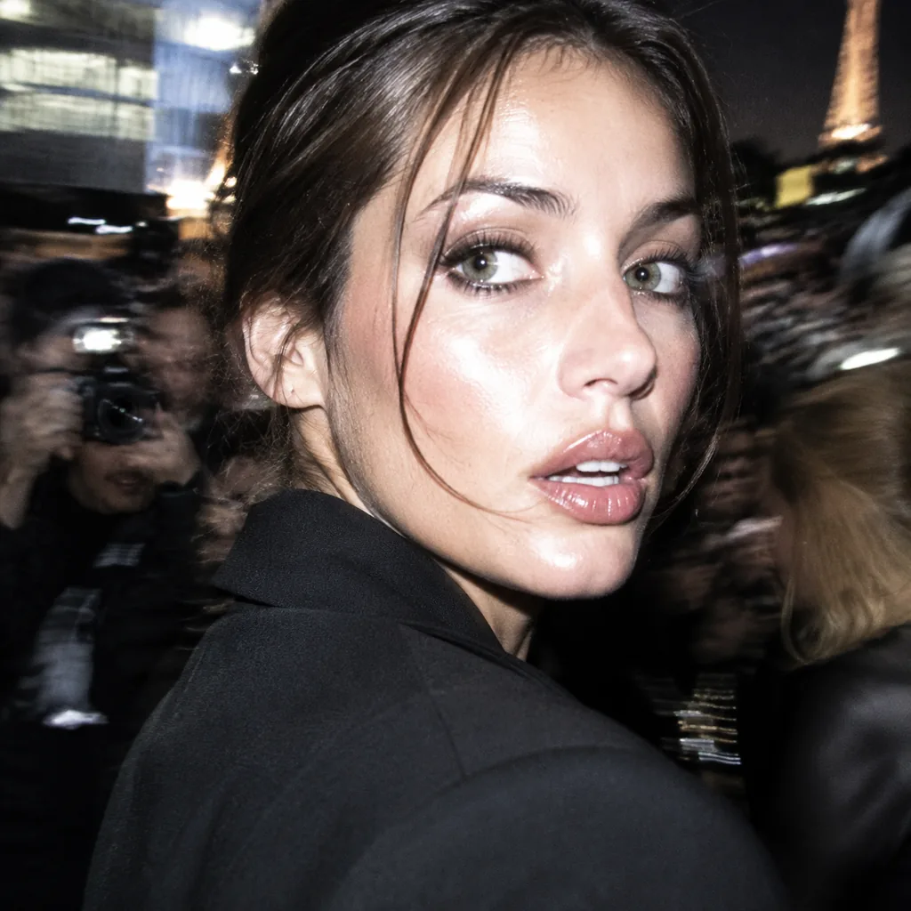 Flash-lit paparazzi close-up of a dark-haired woman in a black blazer glancing back over her shoulder, blurred photographers and the Eiffel Tower behind her at night